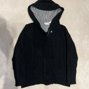 Stickad Kofta  - Säljer en svart stickad hoodie med dragkedja och rutigt foder i huvan. Tröjan har ett snyggt kabelstickat mönster och långa ärmar. Perfekt för kyliga dagar! Vid minsta fråga eller fundering hör av er🤝
