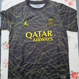 Svart fotbollströja från Paris Saint-Germain - Snygg svart fotbollströja från Paris Saint-Germain med korta ärmar och ett gult geometriskt mönster. Tröjan har klubbens emblem och Jordan-logga på bröstet samt Qatar Airways som sponsor. Perfekt för fotbollsfans!