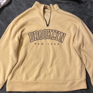 Beige fleece tröja med Brooklyn-tryck - Säljer en mysig beige fleece tröja med texten 'Brooklyn New York' på framsidan. Tröjan har en halv dragkedja vid halsen och långa ärmar. Perfekt för kyliga dagar!