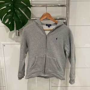 Grå hoodie från Ralph Lauren - Säljer en klassisk grå hoodie från Ralph Lauren med dragkedja och fickor framtill. Den har en broderad logga på bröstet och är tillverkad i mjukt material. Perfekt för en avslappnad stil.