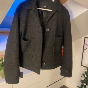 Mörkgrå kappa  - Snygg mörkgrå kappa med stora knappar och två framfickor. Jackan har en klassisk krage och är perfekt för kyligare dagar. Den är stilren och lätt att matcha med olika outfits.