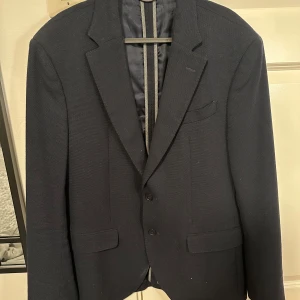 Mörkblå kavaj från Massimo Dutti - Elegant mörkblå kavaj från Massimo Dutti, tillverkad i Portugal med italienskt tyg. Kavajen har en klassisk design med två knappar och fickor fram. Perfekt för en stilren look.
