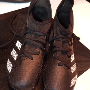 Svarta fotbollsskor från Adidas - Snygga svarta fotbollsskor från Adidas med vita detaljer och snörning. Skorna har en mönstrad yta och dobbar för bra grepp på planen. Perfekta för fotbollsträning och matcher.