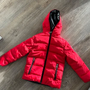 Röd dunjacka från Moncler stl 4 - Snygg röd dunjacka från Moncler med svart foder och logotypdetaljer på insidan av huvan. Jackan har dragkedja och två fickor med dragkedjor framtill. Perfekt för kyliga dagar. stl 4 pris är inte hugget i sten