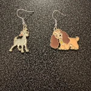 Örhängen med lady och lufsen - Charmiga örhängen med motiv av två olika hundar i silver och bruna nyanser. Perfekta för djurälskare som vill ha något unikt och lekfullt i sin smyckessamling.