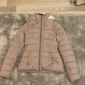 Beige pufferjacka från Hollister - Säljer en snygg beige pufferjacka från Hollister med dragkedja. Jackan har en mysig fodrad huva och långa ärmar. Perfekt för kyliga dagar!