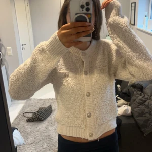 Beige stickad kofta med knappar - Mysig beige stickad kofta med dekorativa knappar framtill. Koftan har en ribbad kant vid ärmslut och nederkant, vilket ger en skön passform. Perfekt för kyligare dagar.