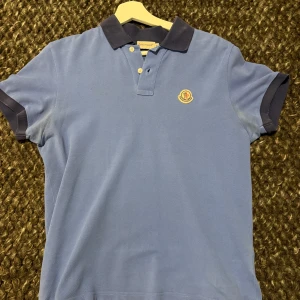 Moncler polo - Säljer en snygg blå pikétröja från Moncler med mörkblå krage och ärmslut. Tröjan har en klassisk passform med korta ärmar och knappar framtill. Perfekt för en stilren look. Den är äkta och köpt några år sen för ungefär 2000. Skriv om du har fler funderingar!