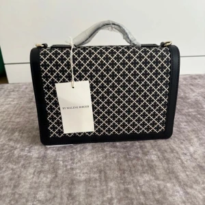 Svart och vit handväska från By Malene Birger - Elegant handväska i svart och vitt med ett stilrent rutmönster. Väskan har en praktisk dragkedja och en avtagbar axelrem. Perfekt för att ge en touch av klass till din outfit.