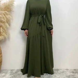 Olivgrön långklänning med knytband - Aldrig använd. Storleken utgår utifrån hur lång man är, 2XL = 172-175 cm. Passar som en vanlig XS/S/M. Elegant olivgrön långklänning med ett avtagbart knytband i midjan. Klänningen har långa ärmar och en asymmetrisk skärning som ger en unik touch. Perfekt för höst och vinter med sitt mjuka och bekväma material.