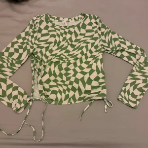 Grön/vit mönstrad topp - Säljer en cool grön och vit topp med ett unikt mönster. Toppen har långa ärmar och snörning på sidorna för en justerbar passform. Perfekt för att ge din outfit en extra touch!