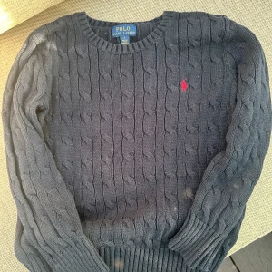Grå stickad tröja från Ralph Lauren - Säljer en stilren grå stickad tröja från Ralph Lauren med kabelstickat mönster. Tröjan har långa ärmar och en liten röd logotyp på bröstet. Perfekt för kyligare dagar.