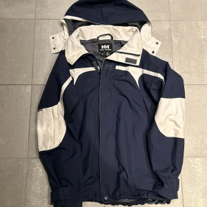 Helly Hansen Jacka - Tja! Säljer nu denna sjyssta Helly Hansen jackan. Den är i väldigt bra skick och passar storlek S. Den har så man kan tajta upp den nertill och huvan går att ta av. Färg i marinblå och vit. Hör av vid frågor!