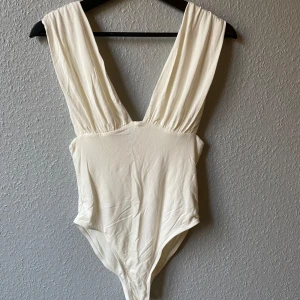 Vit ärmlös bodysuit - Snygg vit bodysuit med djup urringning och breda axelband. Perfekt för en stilren look. Tillverkad i ett mjukt och stretchigt material för en bekväm passform.