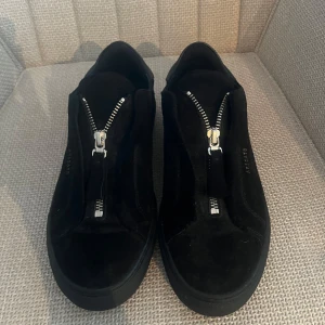 Svarta sneakers från Axel Arigato - Helt oanvända svarta sneakers från Axel Arigato med dragkedja framtill. Ny pris 2500kr. Storlek 39