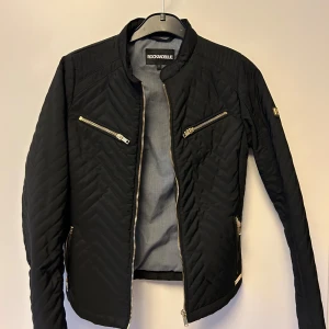 Svart quiltad jacka från Rockandblue - Snygg svart quiltad jacka från Rockandblue med dragkedjor i silverfärg. Jackan har en stilren design med långa ärmar och flera praktiska fickor med dragkedja. Perfekt för en cool och modern look. Nypris 1500kr