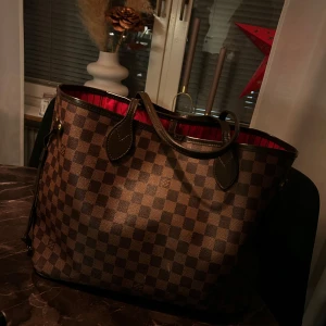 Brun rutig handväska - Snygg brun rutig handväska med röda detaljer på insidan. Väskan har två handtag och en rymlig insida, perfekt för vardagsbruk. Den klassiska rutiga designen ger en tidlös look.