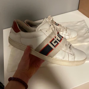 Vita sneakers från Gucci - Snygga vita sneakers från Gucci med röd och blå randdetalj och röd häl. Skorna har snörning och en bred rem med Gucci-logga över vristen. Perfekta för en stilren look. Storlek 42 men passar 43! ✅ dustbags tillkommer vid köp men ingen box. Vid fler frågor och så vidare kom privat! Mvh Levi ✌🏼
