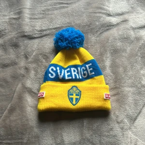 Gul och blå mössa med Sverige-tryck - Snygg gul och blå mössa med texten 'Sverige' och en stor tofs på toppen. Perfekt för att visa ditt stöd för Sverige under kalla dagar.