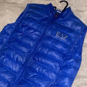 Blå dunväst från Emporio Armani - Snygg blå dunväst från Emporio Armani med dragkedja framtill. Perfekt för kyligare dagar och ger en sportig look. Västen har en broderad logga på bröstet.
