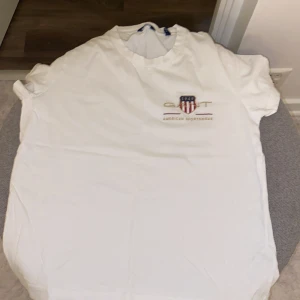 Vit t-shirt från GANT - Säljer en klassisk vit t-shirt från GANT med broderad logga och texten 'American Sportswear' på bröstet. T-shirten har korta ärmar och rund halsringning. Perfekt för en avslappnad stil.