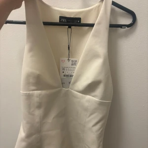 Vit topp från Zara - Elegant vit klänning från Zara med djup v-ringning. Perfekt för en stilren look. Klänningen är ärmlös och har en figurnära passform.