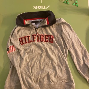 Grå tröja med dragkedja från Tommy Hilfiger - Säljer en grå tröja från Tommy Hilfiger med dragkedja och hög krage. Tröjan har röd text på bröstet och en amerikansk flagga på ärmen. Perfekt för en avslappnad stil.