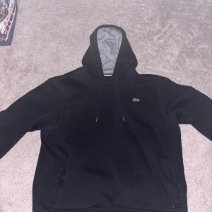 Svart hoodie från Lacoste - Säljer en stilren svart hoodie från Lacoste med grå insida i huvan. Den har en klassisk design med dragsko i huvan och den ikoniska krokodilloggan på bröstet. Perfekt för en avslappnad look.