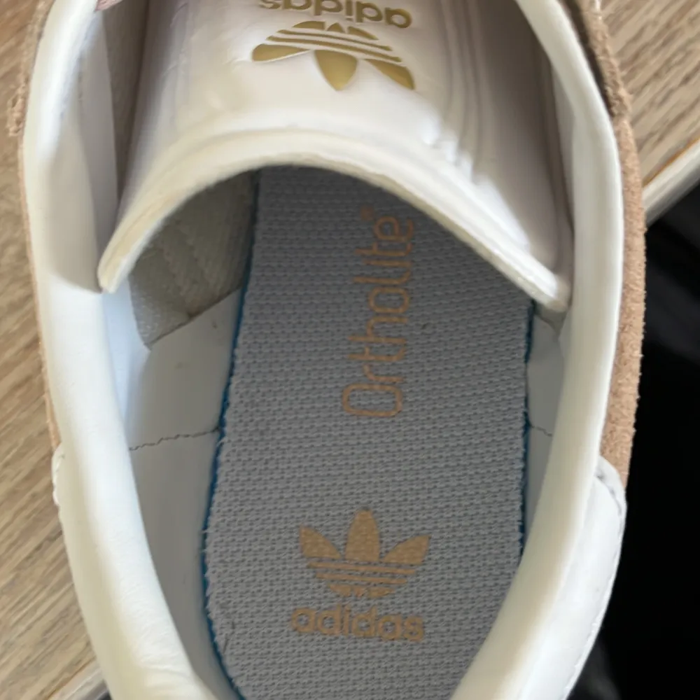 Säljer ett par snygga beige Adidas Gazelle sneakers i mocka. De har de klassiska vita ränderna på sidan och 'GAZELLE' i guldtext. Skorna är i bra skick och perfekta för både vardag och fest. De har en vit sula och snörning framtill.. Kengät.