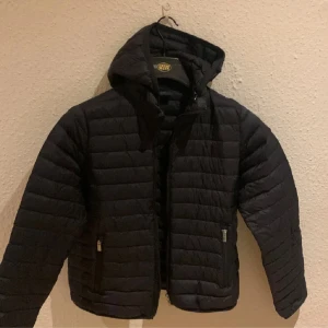 Svart pufferjacka från Peak Performance - Säljer en snygg svart pufferjacka från Peak Performance. Den är perfekt för kyliga dagar med sitt quiltade mönster och dragkedja framtill. Jackan har en huva och praktiska fickor med dragkedjor. Den är i bra skick och redo för nya äventyr!