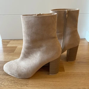 Beige mocka stövletter med klack - Säljer ett par snygga beige stövletter i mocka. De har en rund tå och en stabil klack som ger lite extra höjd. Stövletterna har en praktisk dragkedja på sidan för enkel på- och avtagning. Perfekta för både vardag och fest!