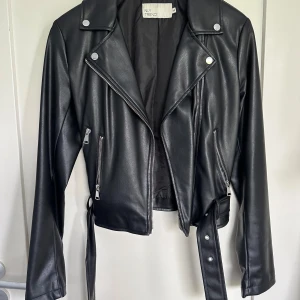 Skinnjacka dam stl 34/xs - Svart skinnjacka i storlek 34/xs från Nelly. Dragkjede stängning. Dragkedje och nitar detaljer, fickor med dragkedja. Bälte som man kan ta bort i midjan. Inga defekter, bara använd ett fåtal gånger. Är som i nyskick.