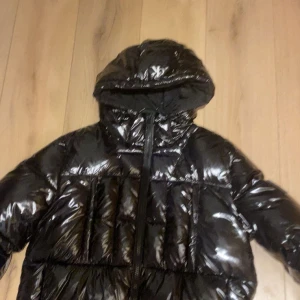 Svart glansig pufferjacka från Calvin Klein Jeans - Säljer en supercool svart glansig pufferjacka från Calvin Klein Jeans. Jackan har en stor huva och dragkedja framtill. Perfekt för att hålla sig varm och stilren under kalla dagar. Den är i nyskick och redo för nya äventyr!