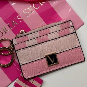 Rosa randig plånbok från Victoria's Secret - Säljer en superfin rosa randig plånbok från Victoria's Secret. Den har en snygg V-detalj på framsidan och kommer med en praktisk dragkedja. Perfekt för att hålla ordning på kort och småsaker. Den är liten och smidig, perfekt för väskan eller fickan!