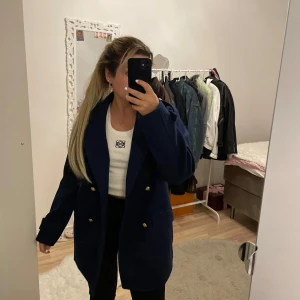 Mörkblå kappa med guldknappar - Säljer en stilren mörkblå kappa med dubbelknäppning och snygga guldknappar. Perfekt för höst och vår, den har en normal passform och långa ärmar. Kappan är i nyskick och passar både till vardags och fest. Oanvänd från Nelly. storlek s