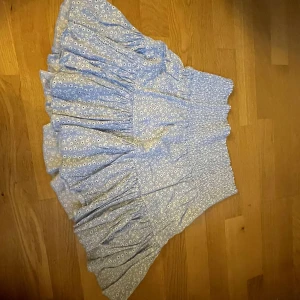 Blå blommig kjol - Säljer en superfin blå kjol med små vita blommor. Den har en elastisk midja som gör den bekväm och lätt att bära. Kjolen är kort och har en volangdetalj som ger den en söt och lekfull look. Perfekt för sommardagar eller en mysig vårutflykt!