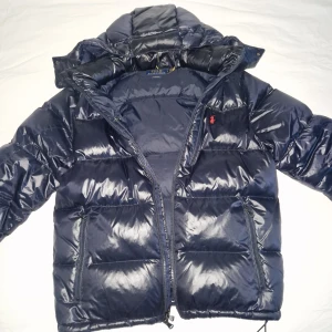 Ralph Lauren Dunjacka  - Stilren dunjacka från Ralph Lauren i färgen glossy navy. Storlek L. Skicket är utmärkt, inga defekter eller liknande. Använts ett par gånger men är för stor för mig, även anledning till varför jag säljer. Ny pris 6k. Är öppen för byten mot  storlek M