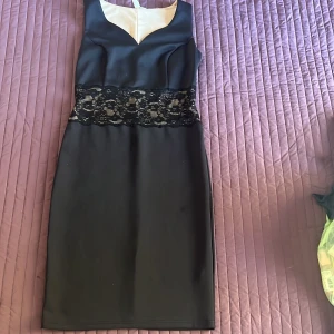 Svart ärmlös klänning med spetsdetalj - Säljer en elegant svart klänning i storlekM. Den är ärmlös och har en vacker spetsdetalj runt midjan som ger en fin kontrast. Klänningen är gjord av polyester och elastan, vilket gör den både bekväm och stretchig. Perfekt för en festlig kväll eller
