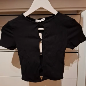 Svart ribbad topp från FB Sister - Säljer en helt ny svart ribbad topp från FB Sister i storlek XXS. Toppen har en unik design med öppningar framtill. Perfekt för en stilren & trendig look! Passar även upp till S då den är elastisk.
