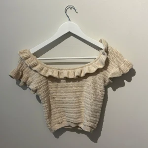 Beige stickad offshoulder topp från H&M - Säljer en söt beige stickad offshoulder topp från H&M. Toppen har volangdetaljer runt halsen ochärmarna och är perfekt för en somrig look. Den är bekväm och stilren.