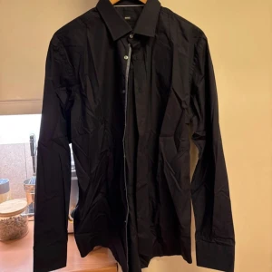 Svart skjorta från Boss - Snygg svart skjorta från Boss i slim fit med långa ärmar och klassisk krage. Skjortan har knappar framtill och vid ärmsluten, vilket ger en stilren look. Perfekt för en elegant stil.
