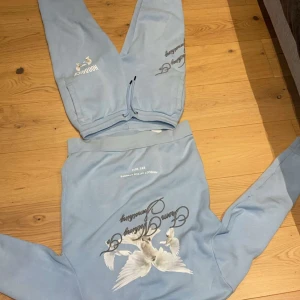 Ljusblå hoodie och byxor från Hoodrich - Säljer ett set med ljusblå hoodie och matchande byxor från Hoodrich. Hoodien har ett tryck med text och en fågel på framsidan. Byxorna har dragsko i midjan och texttryck på benet. Perfekt för en avslappnad stil. Pris kan diskuteras vid en snabb affär!