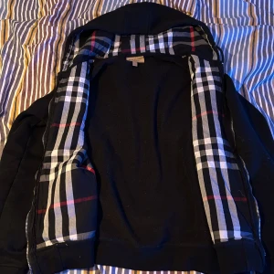 Svart hoodie från Burberry storlek S - Säljer en stilren svart hoodie från Burberry med klassiskt rutigt mönster på insidan av huvan. Jackan har dragkedja och är tillverkad i bomull med elastan för extra komfort. Perfekt för en casual look med en touch av lyx. Inga defekter, säljer för den tyvärr inte passar längre. Skriv för fler frågor, priset kan diskuteras.