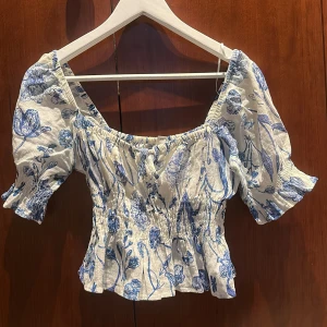 Blommig offshoulder topp - Säljer en söt offshoulder topp från Zara med blått blommönster på vit bakgrund. Toppen har korta puffärmar och en smockad midja för en fin passform. Perfekt för varma dagar!