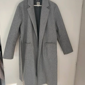 Grå kappa  - Snygg grå kappa från Only med knytskärp i midjan. Den har en klassisk design med långa ärmar och två fickor framtill. Perfekt för kyligare dagar.
