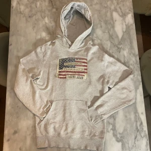 Grå Ralph Lauren hoodie med amerikansk flagga  - Skick: 8/10. Hoodien är i bra skick, ett litet hål vid hörnet av flaggan men inget märkvärdigt då det är en ”sliten” design (se bild 4).   Märke: Ralph Lauren   Storlek: M  Färg: Grå ✅Tar även byten på allt 