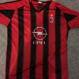 Retro Milan tröja - Säljer en klassisk röd och svart randig fotbollströja från Adidas med korta ärmar. Tröjan har ett broderat klubbmärke på bröstet och Opel som sponsorlogga. Perfekt för fotbollsfans som vill visa sitt stöd. Oversized