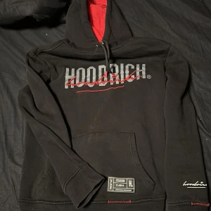 Svart hoodie från Hoodrich - Säljer en svart hoodie från Hoodrich med röd insida av huvan och tryck i vitt och rött på både fram- och baksidan. Hoodien har en stor ficka framtill och ribbade muddar vid ärmslut och nederkant. Perfekt för en casual look.