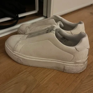 Vita sneakers - Säljer ett par vita sneakers med en stilren design och tjock sula. Skorna har en enkel slip-on stil utan snörning, vilket gör dem lätta att ta på och av. Perfekta för en avslappnad look. Tvättas innan den skickas 