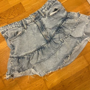 Blå jeanskjol från Gina  - Snygg blå jeanskjol med volangdetaljer och fransig kant. Perfekt för en avslappnad stil med en touch av edge. Kjolen har en klassisk jeanslook med bälteshällor och knappstängning.